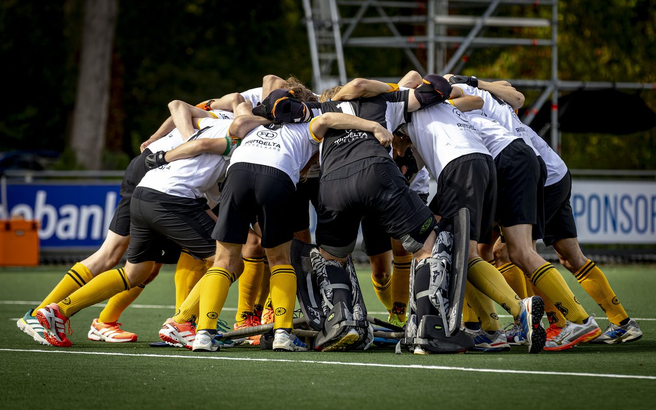 Hockeyteam in wit en zwart met gele sokken staat in een kring op het veld, armen om elkaar heen