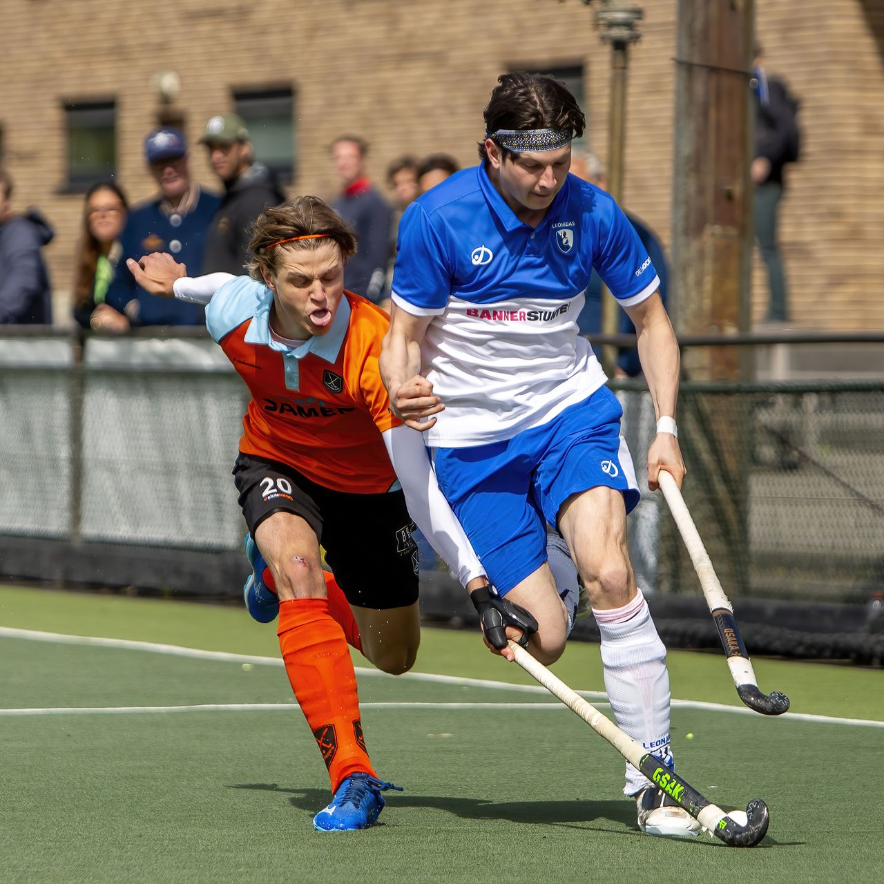 Hockeyspeler in blauw drijft de bal op terwijl een tegenstander in oranje hem met open mond probeert bij te houden