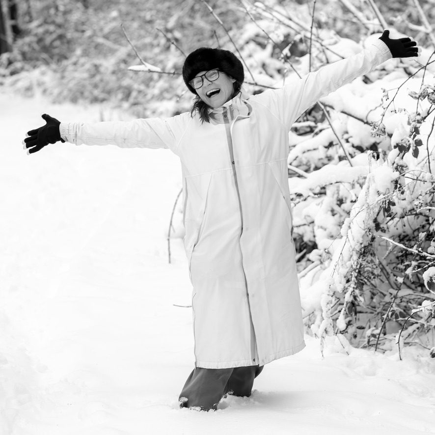 Vrouw in witte jas staat met gespreide armen lachend in de sneeuw, in zwart-wit