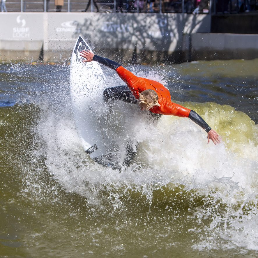 Surfer in oranje pak maakt een draai in een golfslagbad, het water spat op